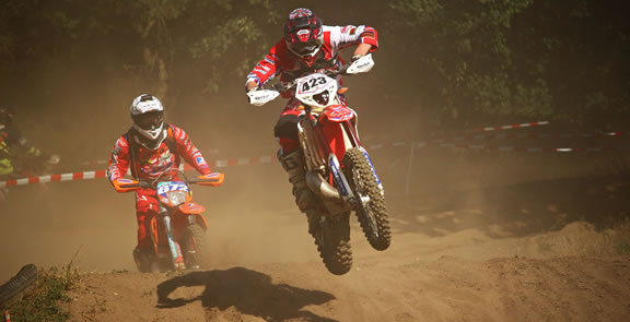 Motocross riders mid-race with one airborne over a jump and another close behind on a dirt track